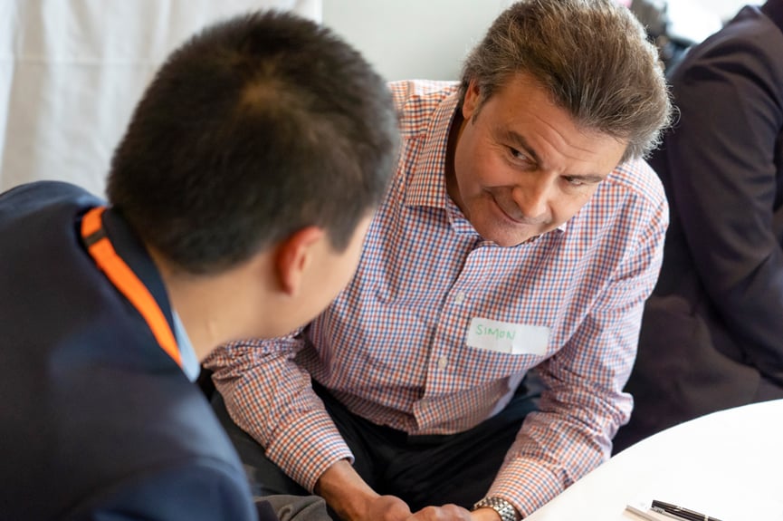 A male mentor wearing a checkered shirt is looking kindly at a male student. The student has his back to the camera.